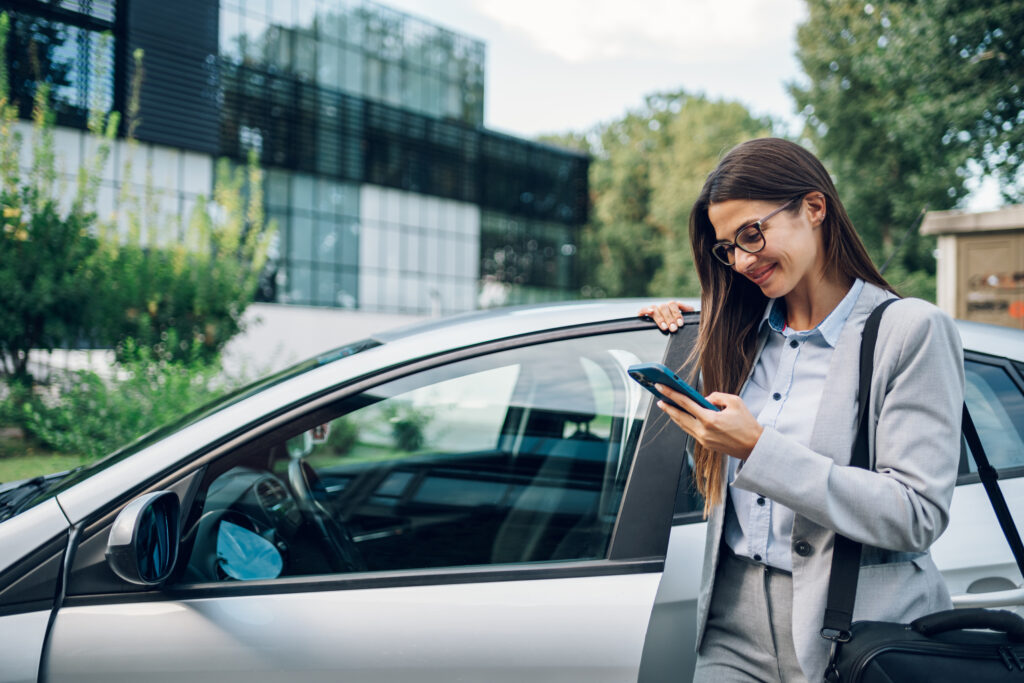 Woman in a blazer stands beside an open car door, looking at her smartphone outdoors near a modern glass building.